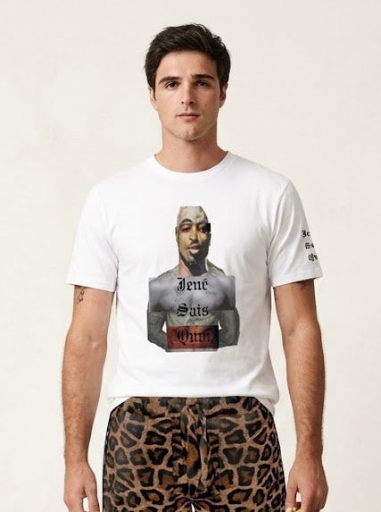 A male model stands against a light beige studio background wearing a white graphic T-shirt, leopard-print boxer briefs, leopard-print socks, and sandals, while holding a green and yellow patterned cap.